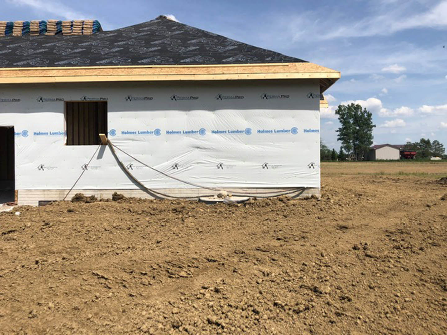 Partially built house with black roof, exposed framing, white tarp covering dirt field, stack of construction bags, and leafy tree nearby