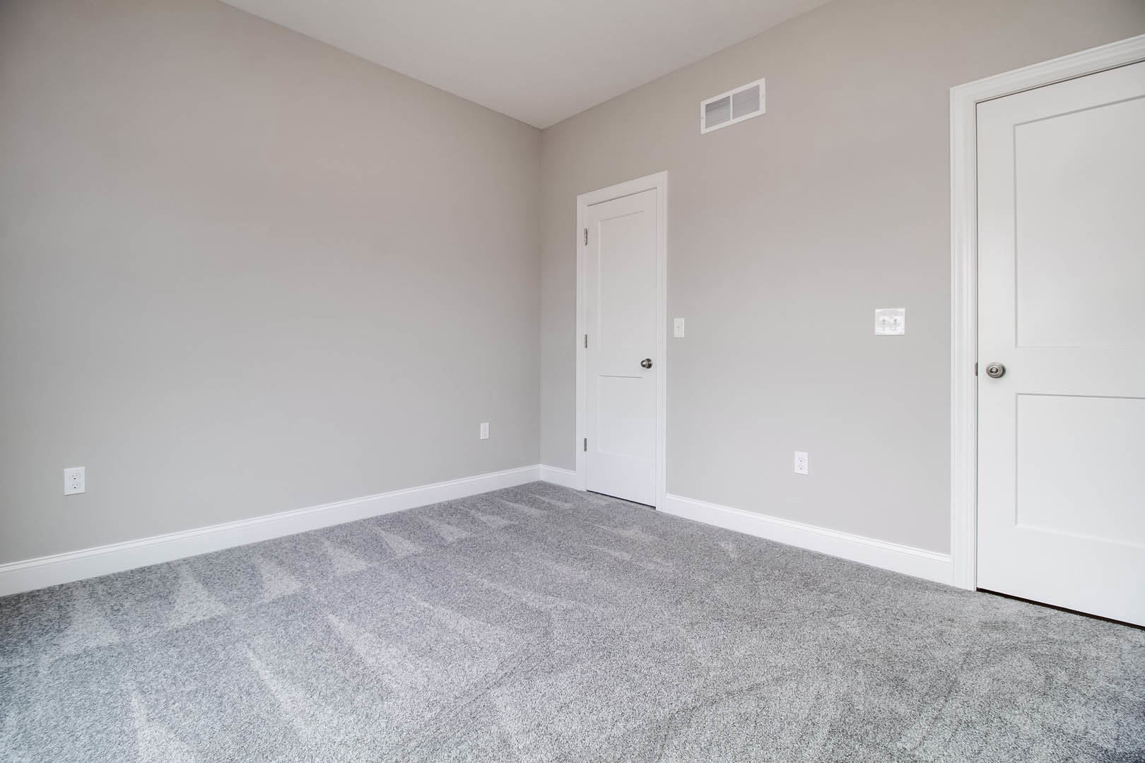 Corner of a room with white walls, a white door featuring a silver knob, grey carpet flooring, and a white rectangular vent near the baseboard.