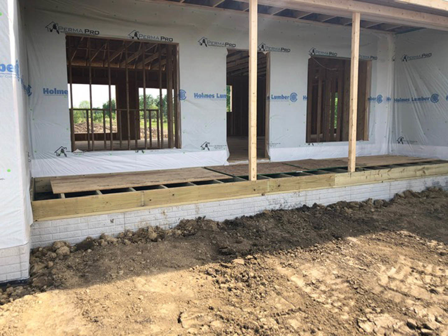 Wood-framed house under construction with exposed beams, brick walls, and dirt-covered ground; unfinished windows visible on exterior.