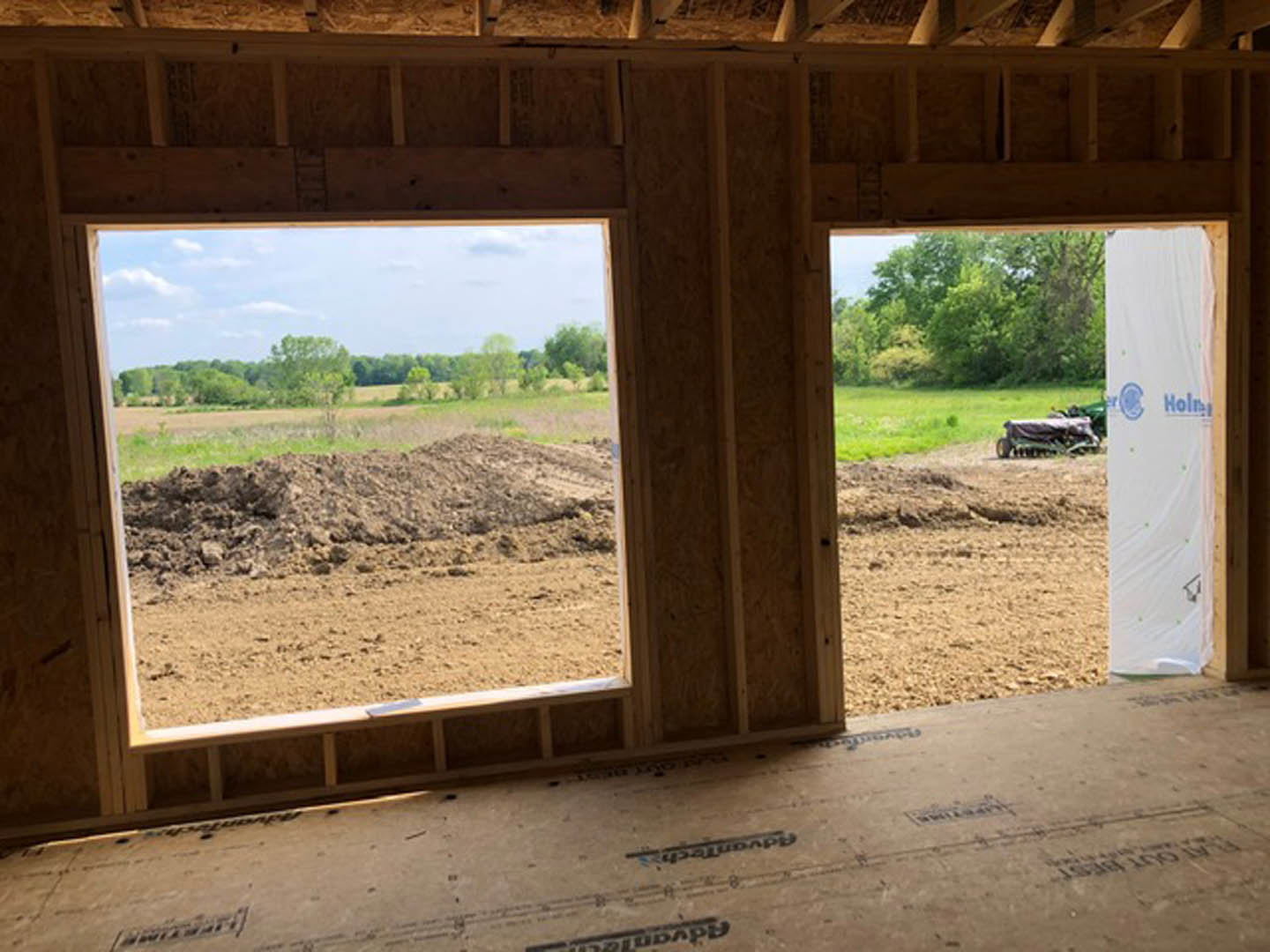Large rectangular window framed in unfinished wood overlooks dirt construction site with grass and trees in background; white sign with blue text visible near window.