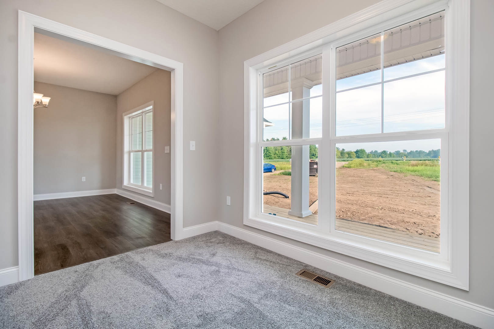 Spacious room featuring a large window overlooking a dirt field, hardwood flooring with a metal floor vent, white plaster walls, and simple molding.