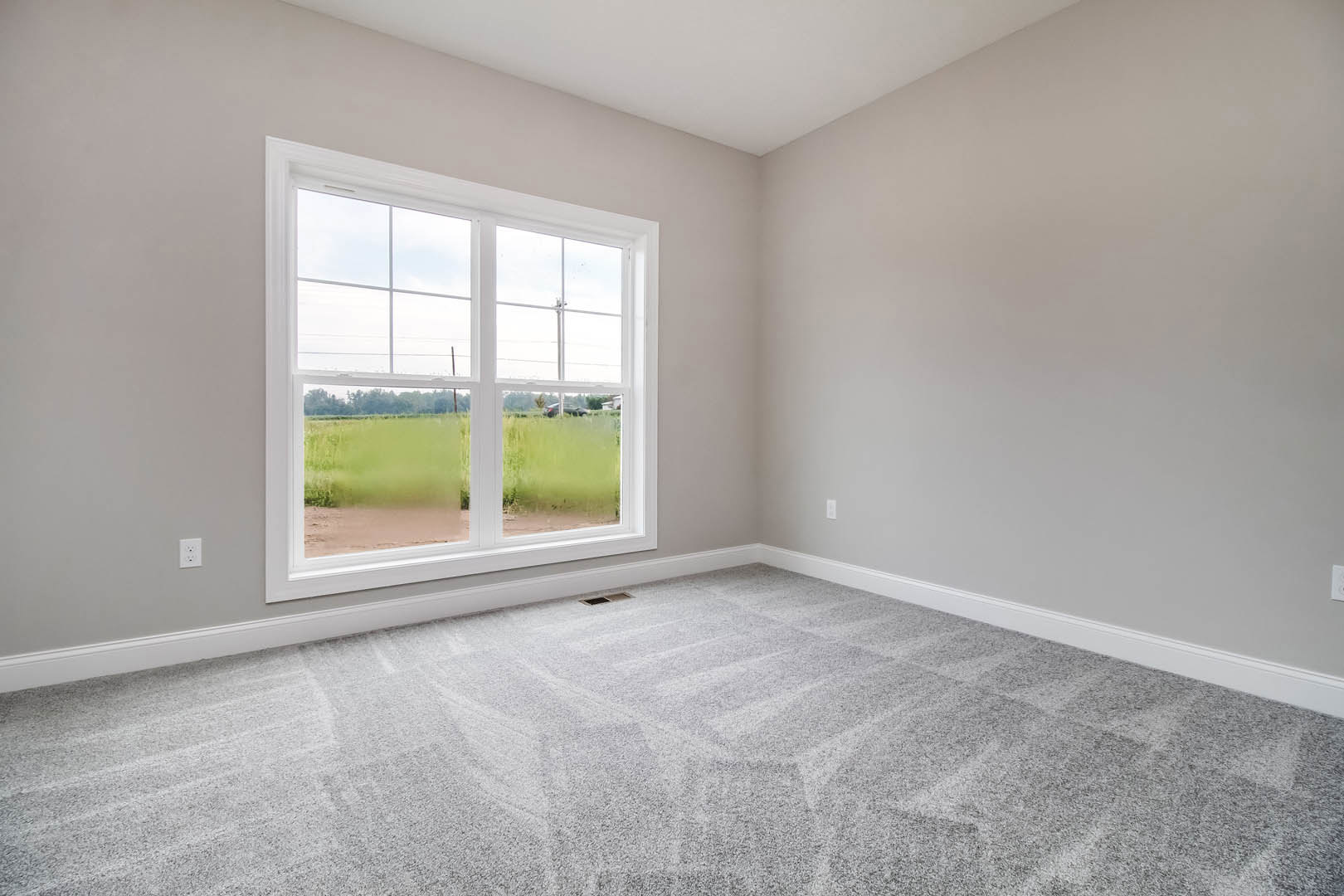 Bright room with white walls, grey carpet flooring, large window overlooking green grass and parked car, white ceiling