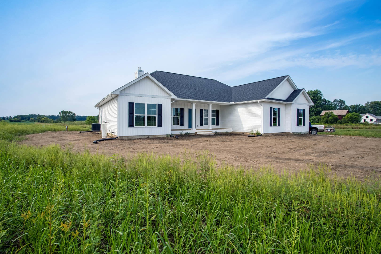White house with blue roof set on a dirt and grass field, Southfork Ranch visible in the background, large windows and clear blue sky overhead