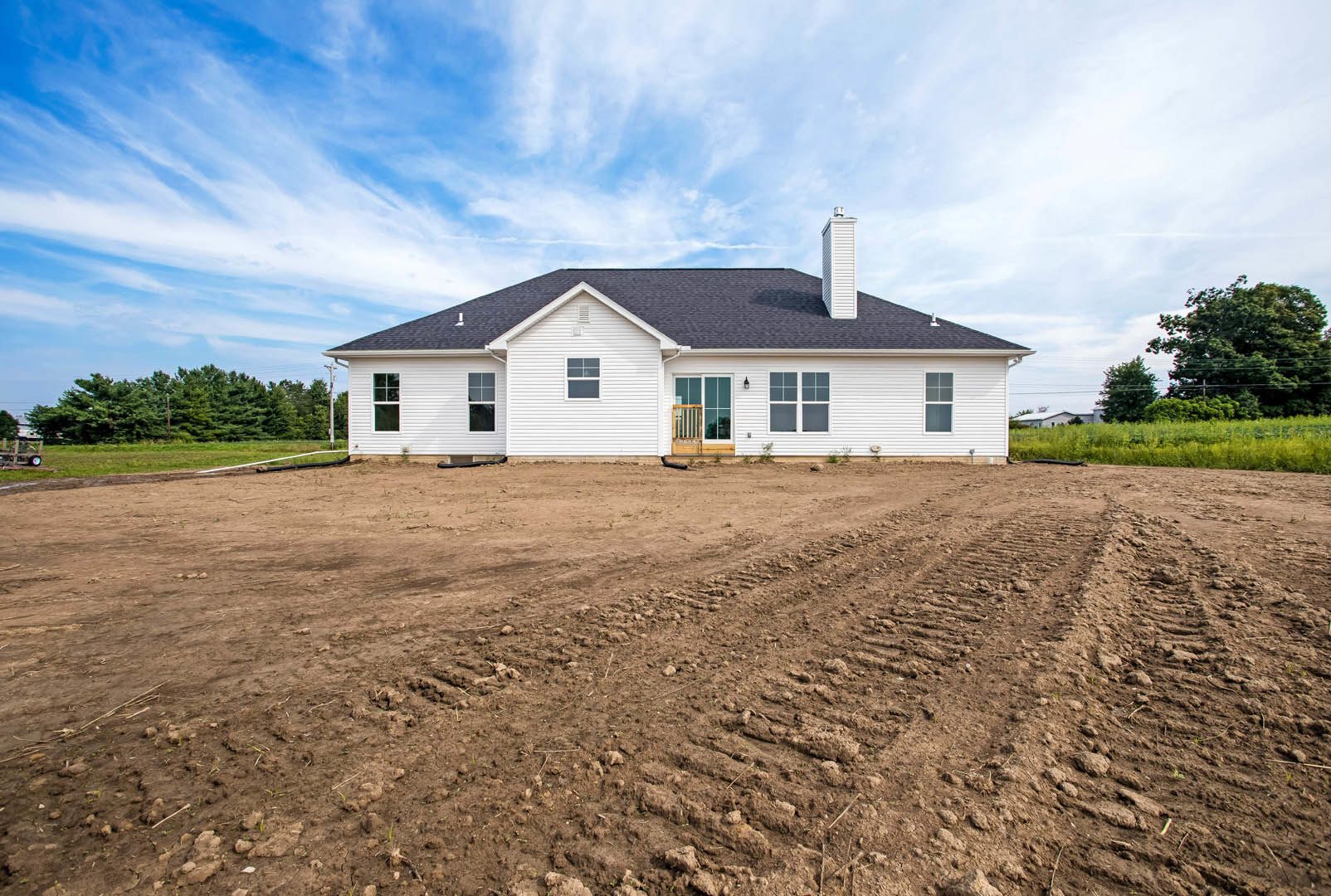 White farmhouse with wooden front door, dirt lot with tire tracks, chimney, and surrounding trees under cloudy sky