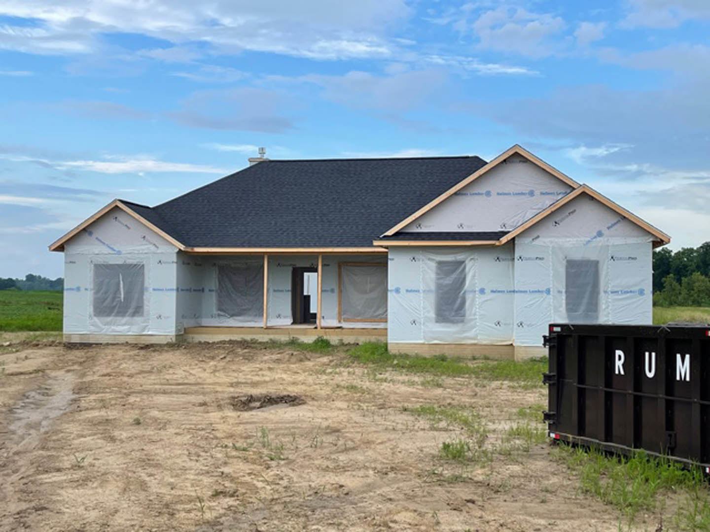Partially built house with exposed framing and plastic sheeting, black dumpster with white lettering in front, dirt yard with patches of grass, white wall visible, cloudy sky