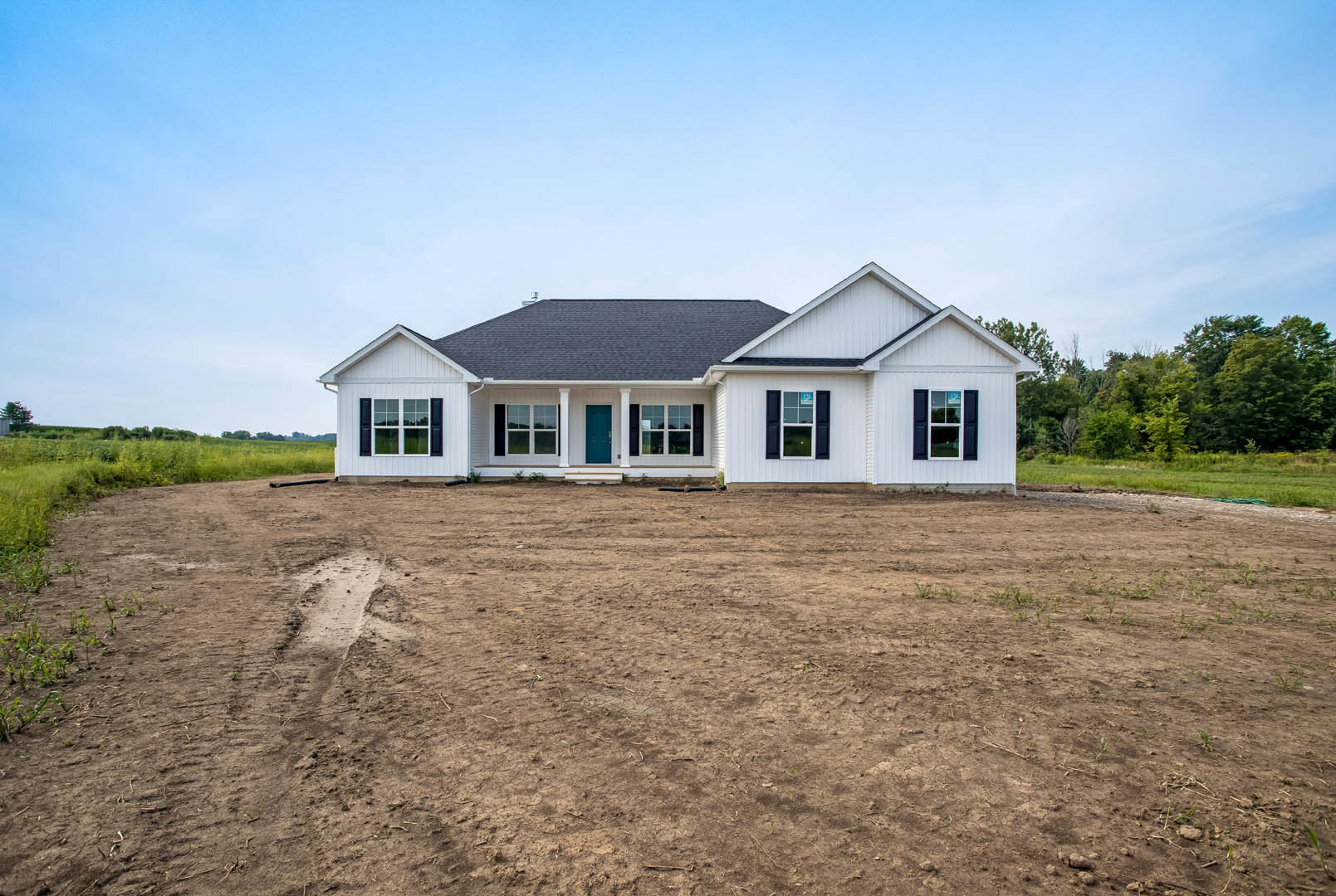 White house with black roof and blue front door, surrounded by a dirt field, grassy patches, and sparse trees under a cloudy sky