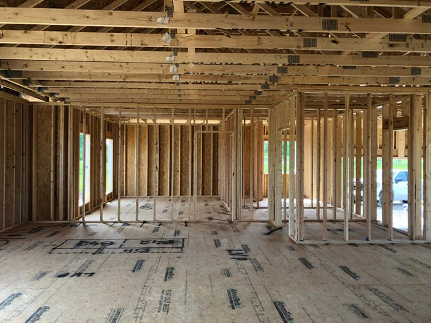Room under construction with exposed wood framing, ceiling beams, unfinished floor marked with black writing, and a window letting in natural light
