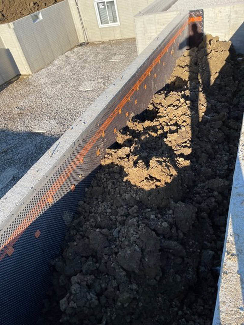 Exposed soil and concrete foundation in a trench beside a white exterior wall with a window, showing construction materials and unfinished ground.