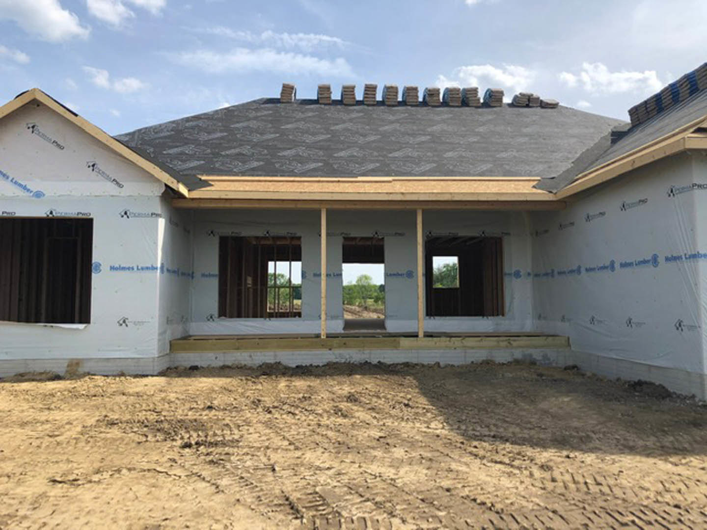 Partially built house with exposed framing and roof, surrounded by dirt road and construction materials, cloudy sky overhead, forest visible in background