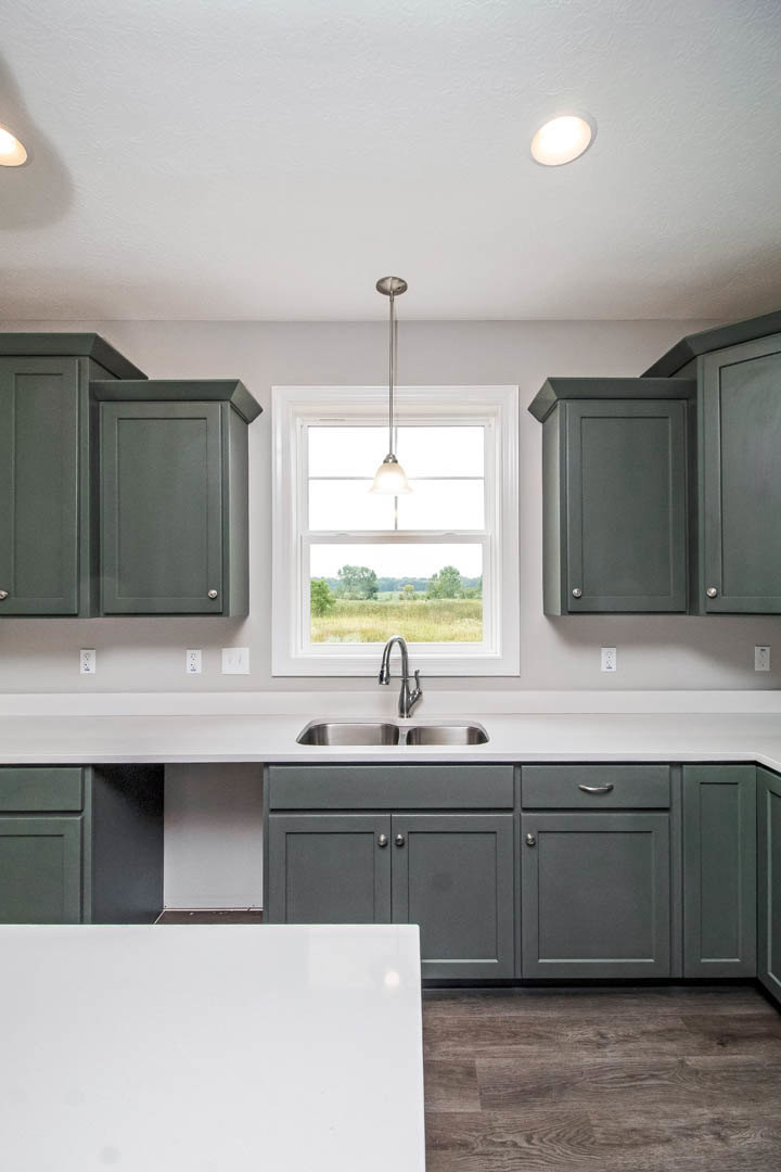 Grey kitchen cabinets with brushed metal hardware, white quartz countertop, stainless steel sink and faucet, wood flooring with black baseboards, large white-framed window above