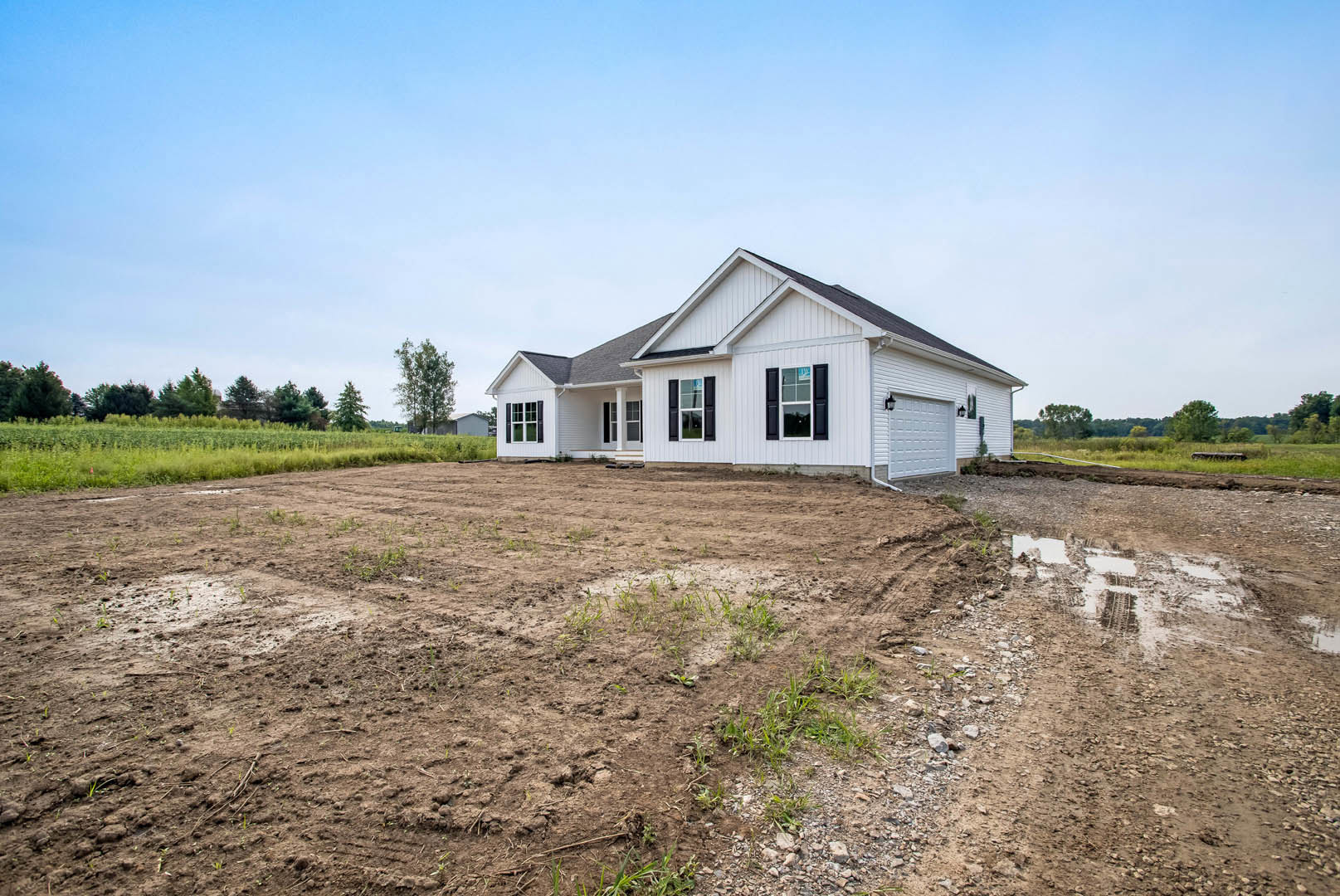 White house with black shutters and white-framed windows, surrounded by a dirt patch with sparse grass, leafy tree nearby, blue sky overhead