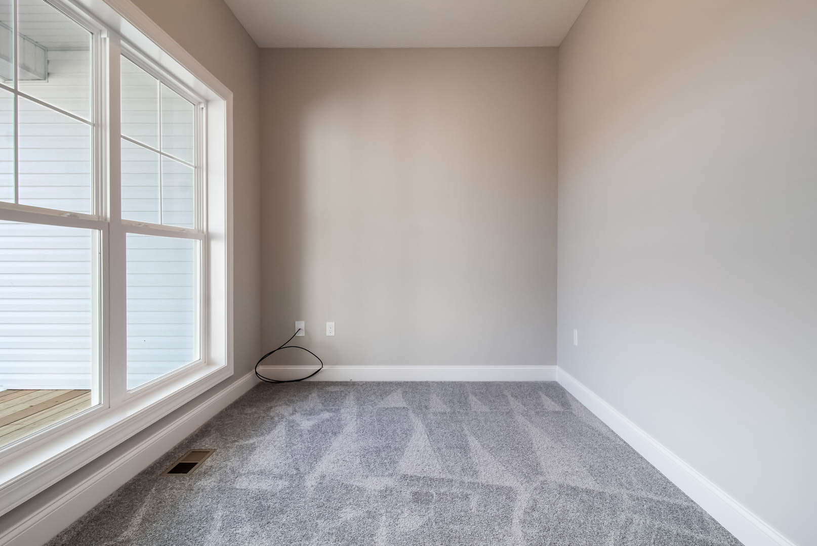 Grey carpeted floor with a black cord near a white wall, large window with white frame letting in natural light, cloudy sky visible outside