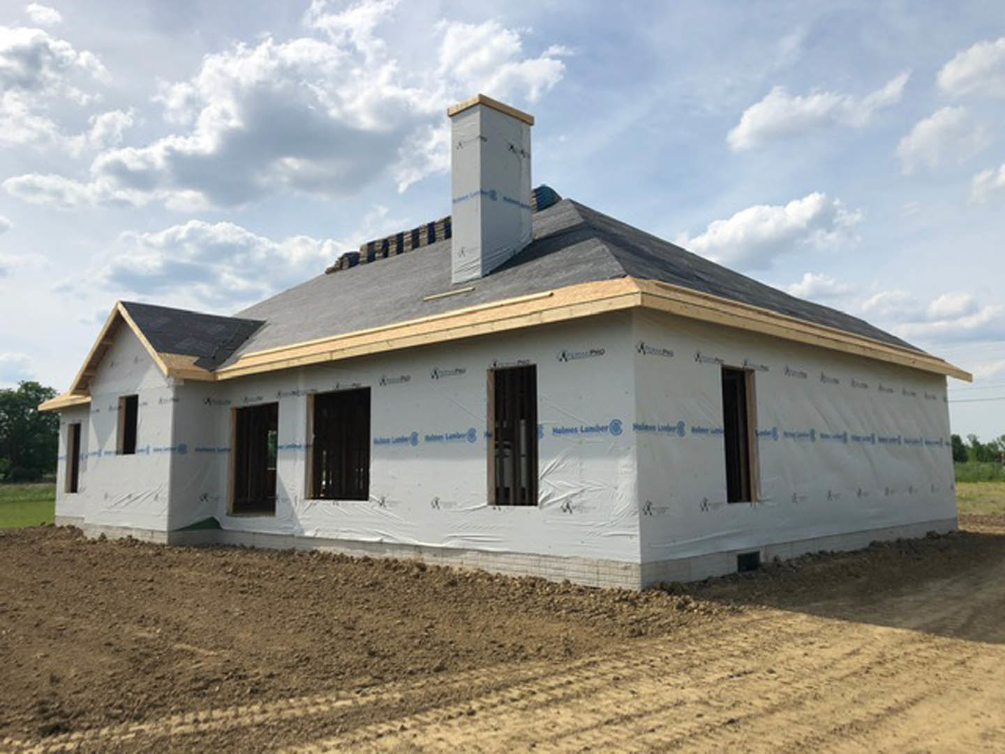 Partially built house with exposed framing, chimney, dirt lot with tire tracks, cloudy sky overhead