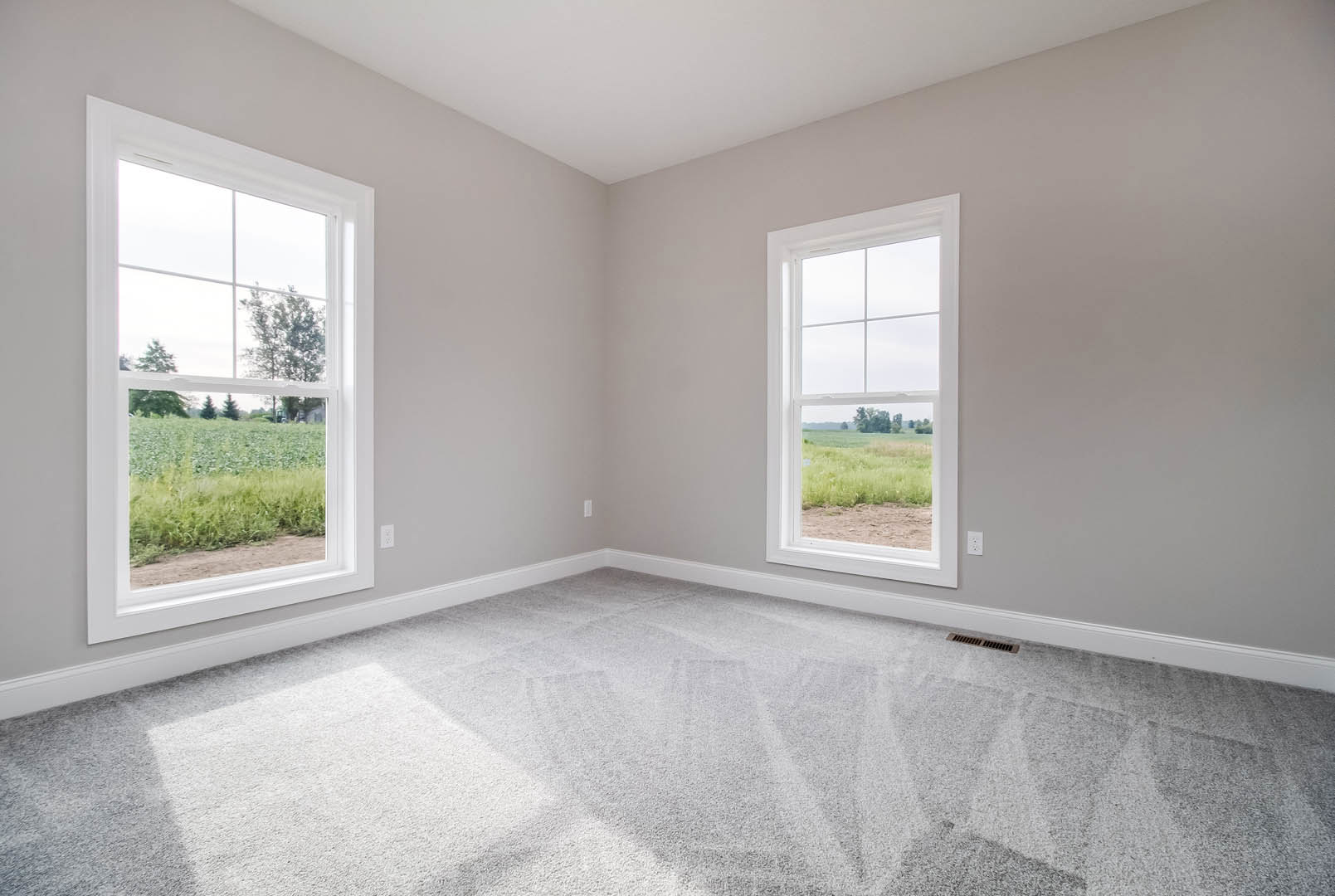 Carpeted room corner with large window overlooking green field and snowy trees, white ceiling, neutral walls
