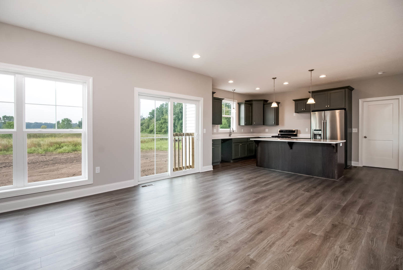 Open kitchen and living room with hardwood floors, white cabinetry, black countertops, sliding glass door overlooking a field, window with view of dirt road, white door with black