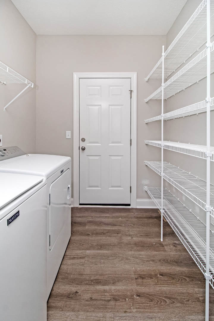 Laundry room with white open shelves, front-loading dryer, wood flooring, and white door with silver handle