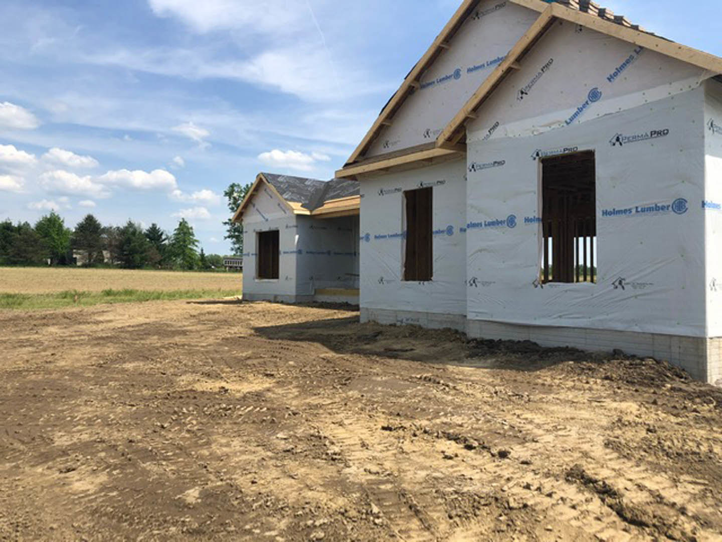 Wood-framed house under construction with exposed plywood, dirt lot in foreground, and leafy trees in background under blue sky