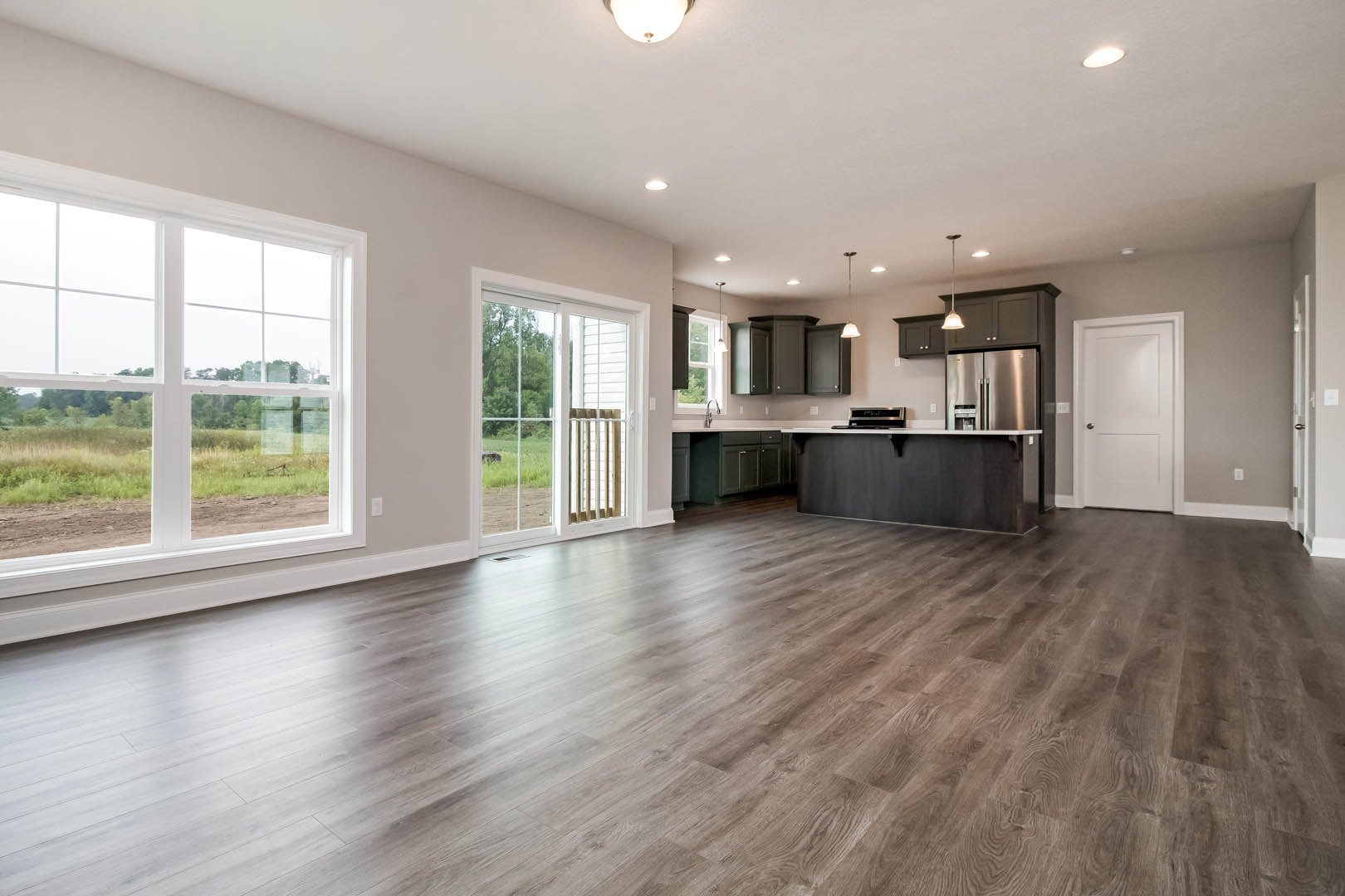 Open kitchen with black cabinetry, white countertops, hardwood flooring, white door with silver handle, large window overlooking grassy field.