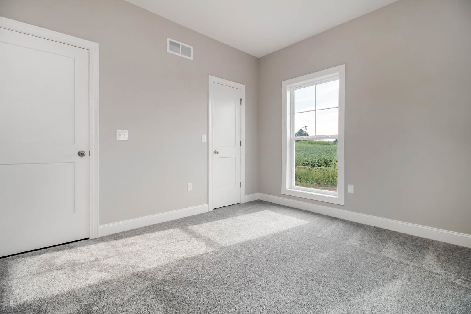 Carpeted room with two white doors featuring silver knobs, rectangular window with white frame overlooking a field, white plaster walls