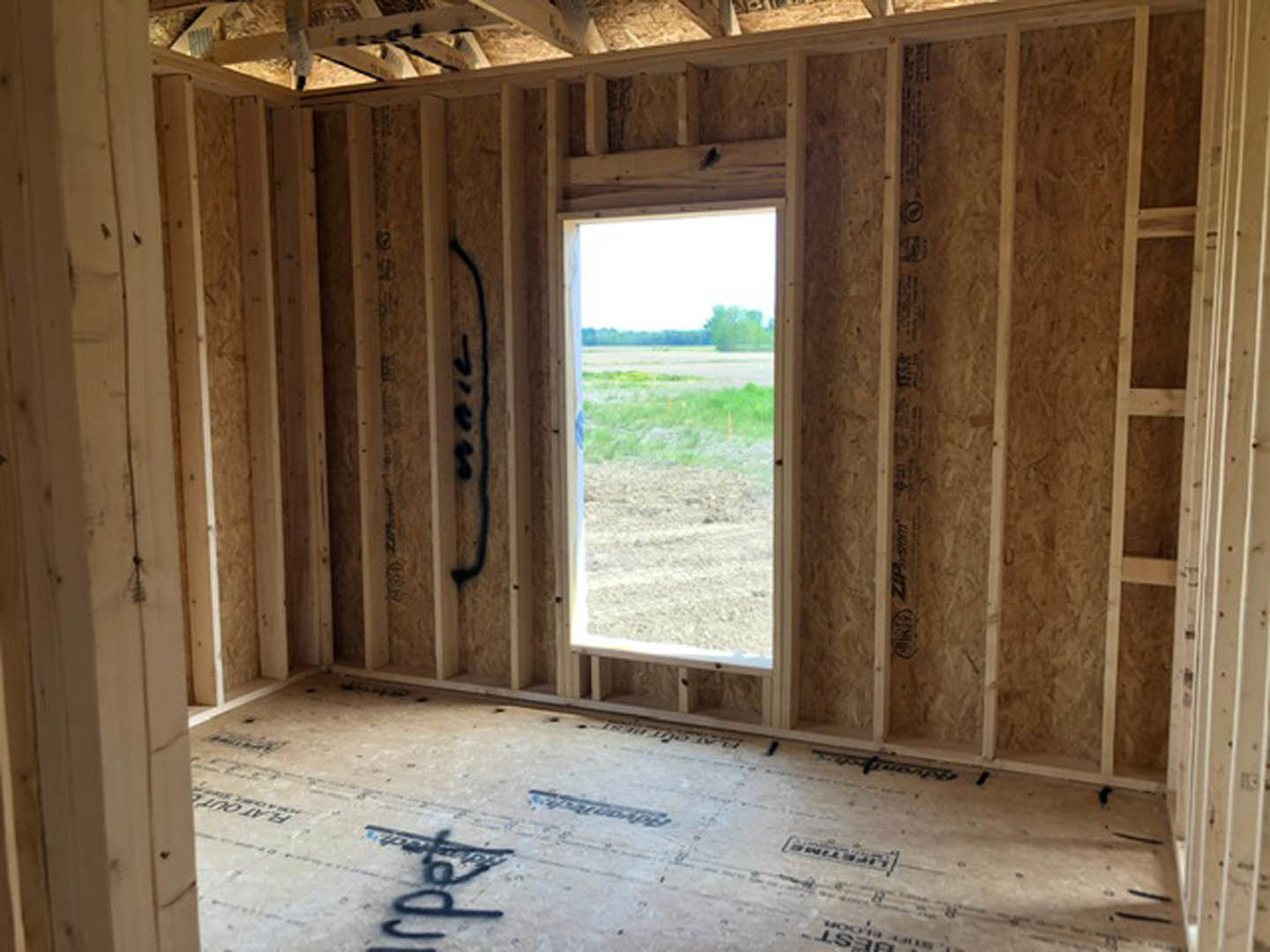 Sunlit room with wood plank flooring, large window overlooking grassy field and trees, open door leading outside, neutral walls
