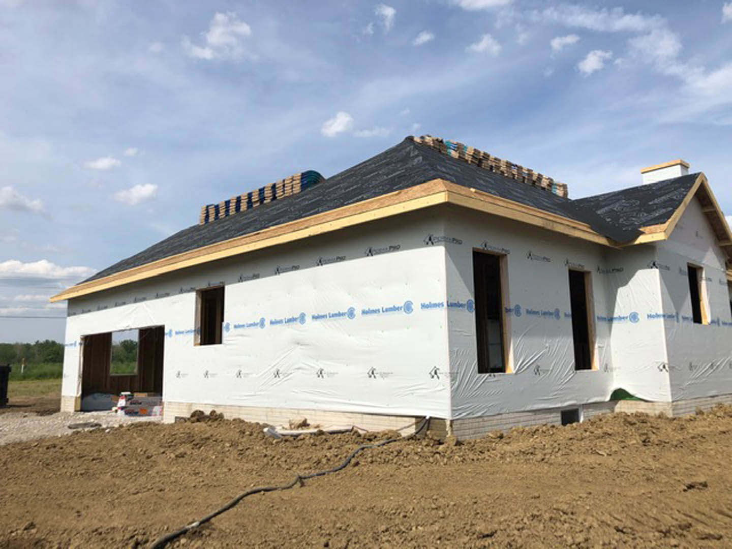 Partially built house with exposed framing and roof, surrounded by dirt lot under blue sky with scattered clouds