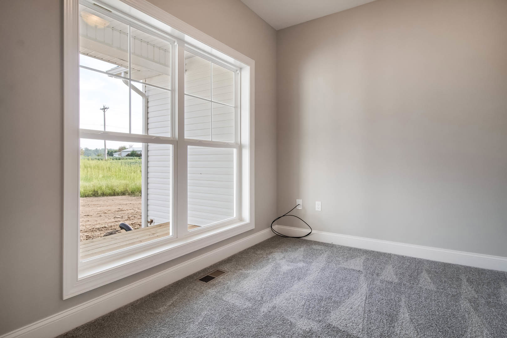 Large window with white trim overlooking a field of long grass, grey carpeted floor with a vent in the corner, black wire running along the wall