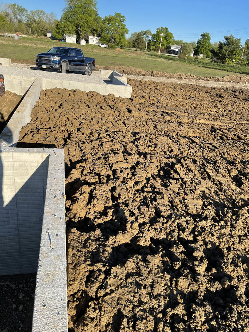 Black pickup truck parked on gravel driveway beside unfinished concrete and brick walls, surrounded by dirt, rocks, and sparse grass with trees in the background.