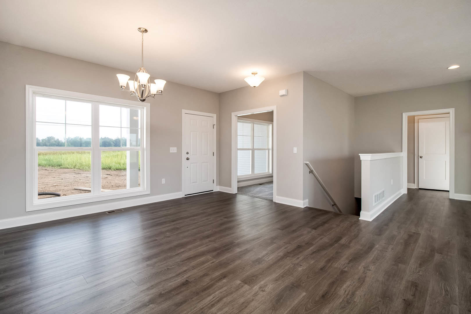 Open living space with hardwood flooring, white walls, modern chandelier, large window overlooking a field, white door with silver handle, and stair rail detail