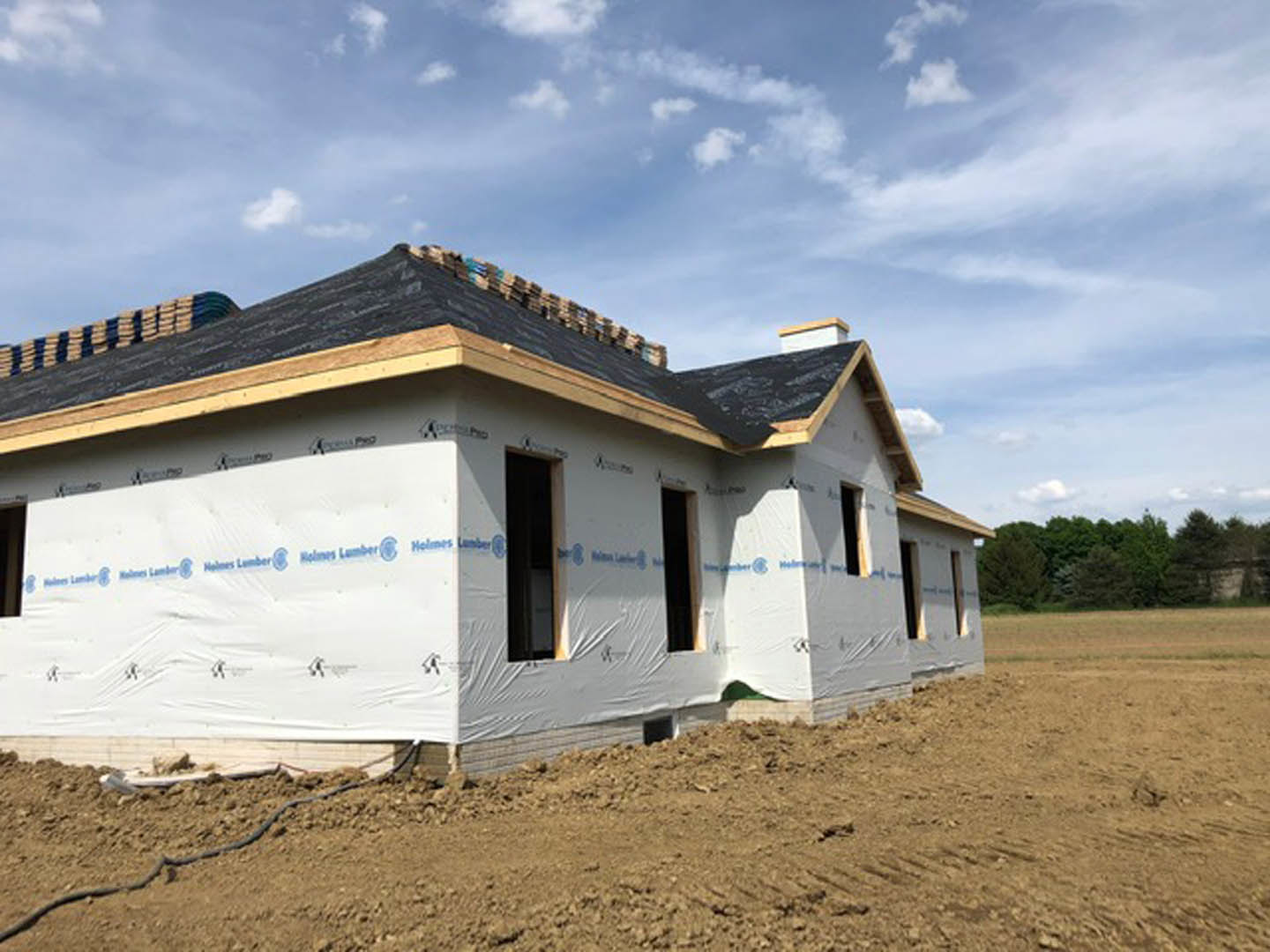 Partially built house with black roof and tarp covering, surrounded by grassy field, trees in background, blue sky with scattered clouds