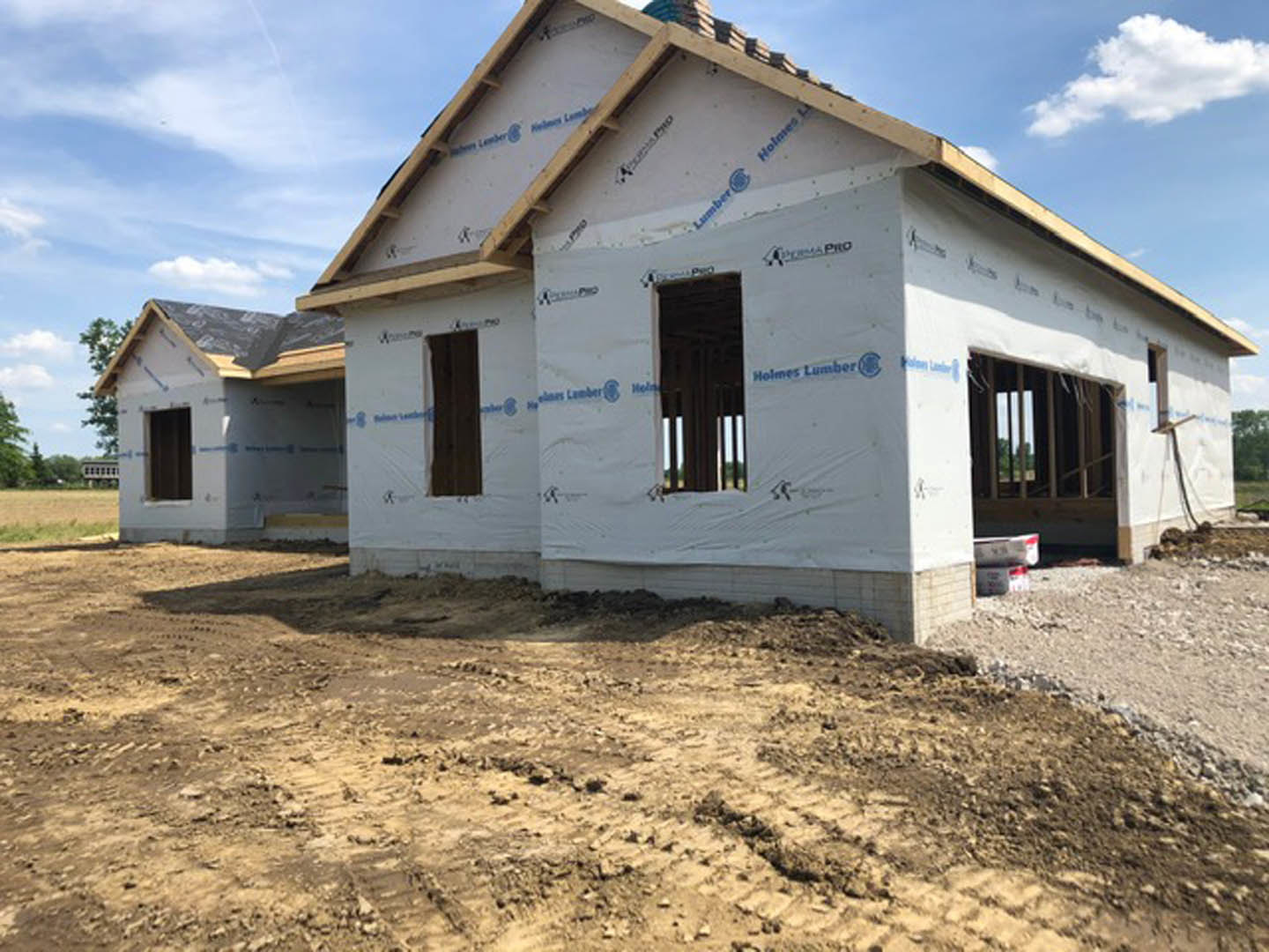 Wood-framed house under construction with exposed beams, dirt foreground, white tarp, and blue sky background
