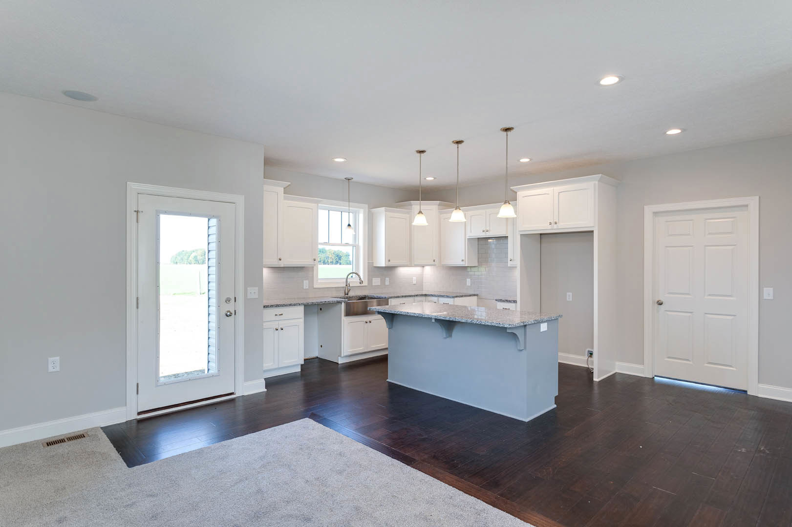 Open-concept kitchen featuring white cabinetry, a central bar with stools, patterned rug on hardwood floor, quartz countertops, stainless steel sink, white-framed window, and a