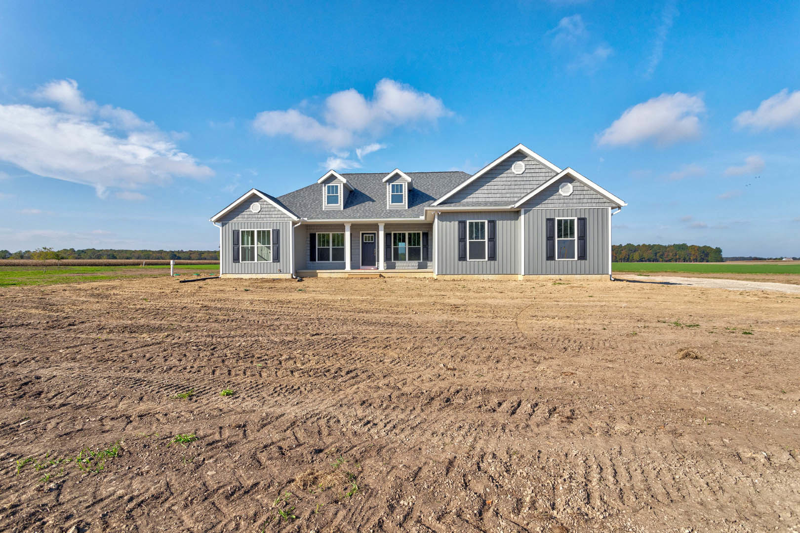 Two-story custom home with white-framed windows, surrounded by a dirt field with visible tire tracks, under a blue sky with scattered clouds