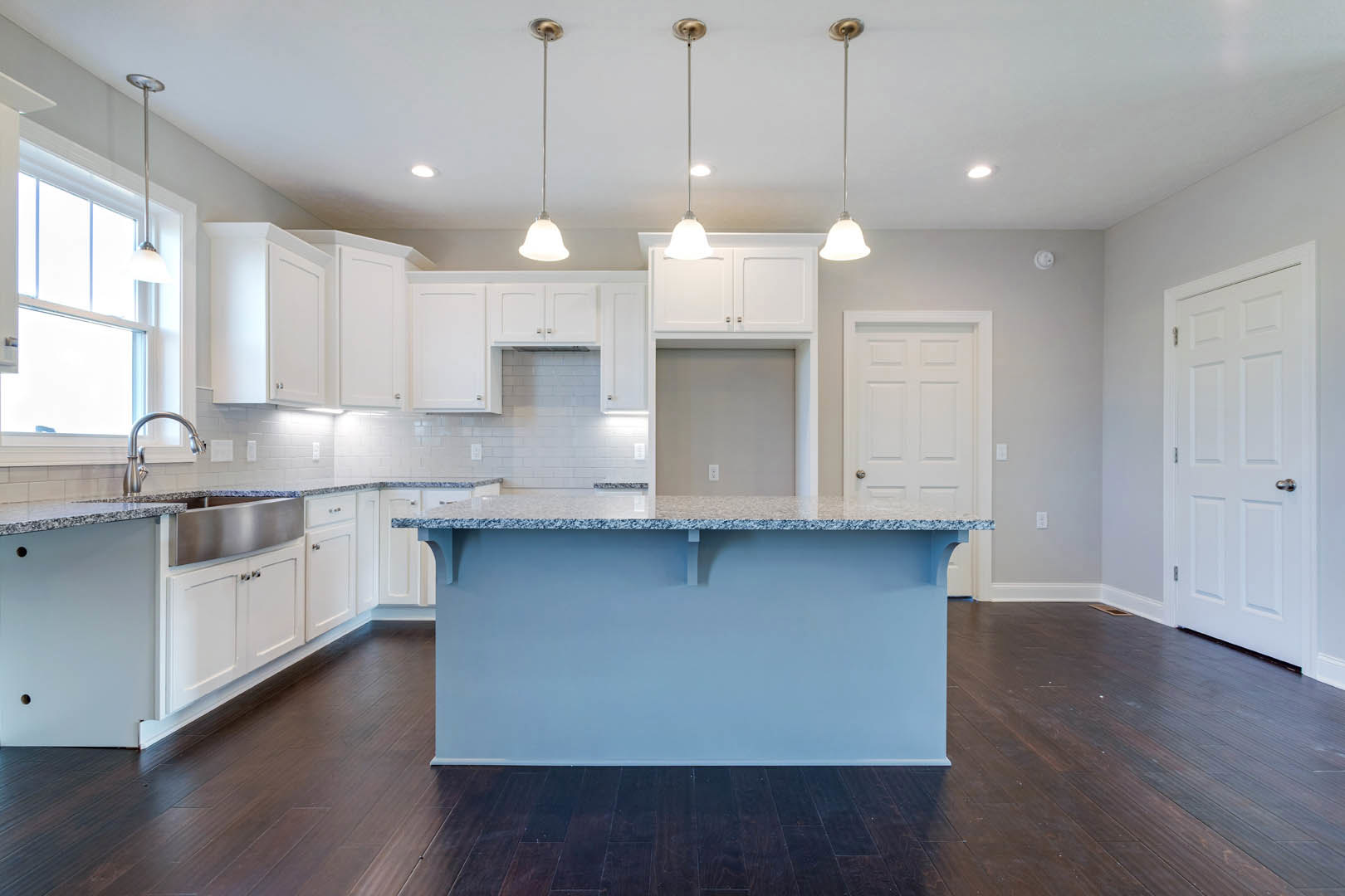 Kitchen with white shaker cabinets, blue countertop bar, stainless steel sink, dark wood flooring, white walls, and a slim metal support pole.