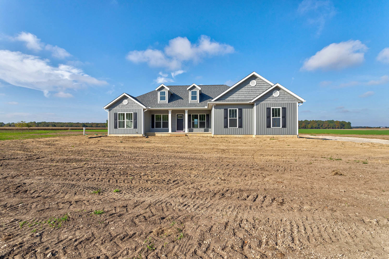 Single-story house with white-framed windows, front porch, and door, surrounded by a dirt field under a blue sky with scattered clouds