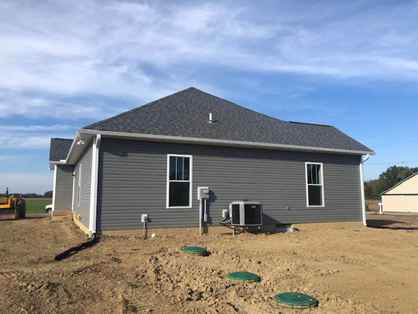 Partially built house with exposed framing, white window frame, green tarp covering dirt, and heater visible under clear blue sky