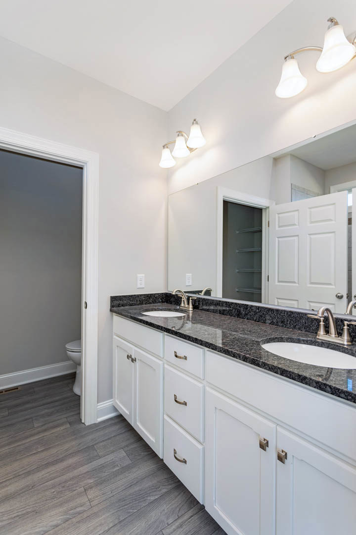 Bathroom with marble countertop, undermount sink, chrome faucet, white cabinetry, grey painted walls, and toilet adjacent to a white door frame