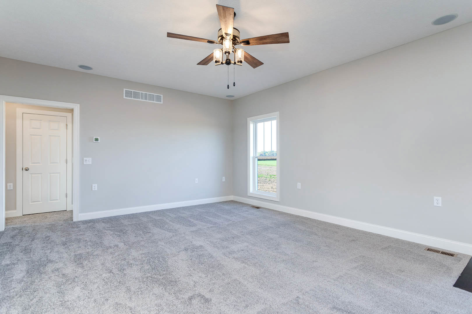 Carpeted room with white walls, ceiling fan with light fixture, white door with silver handle, window overlooking grassy field
