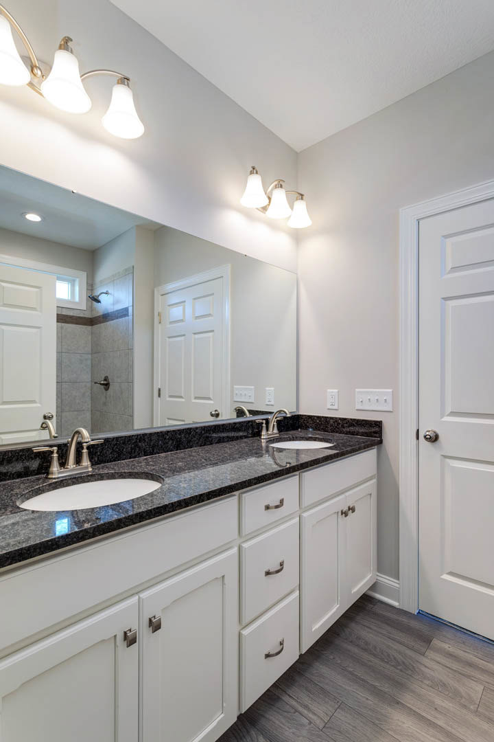 Bathroom with a wide mirror above double sinks, white cabinetry, chrome faucets, and tiled walls; white door with metal handle visible.
