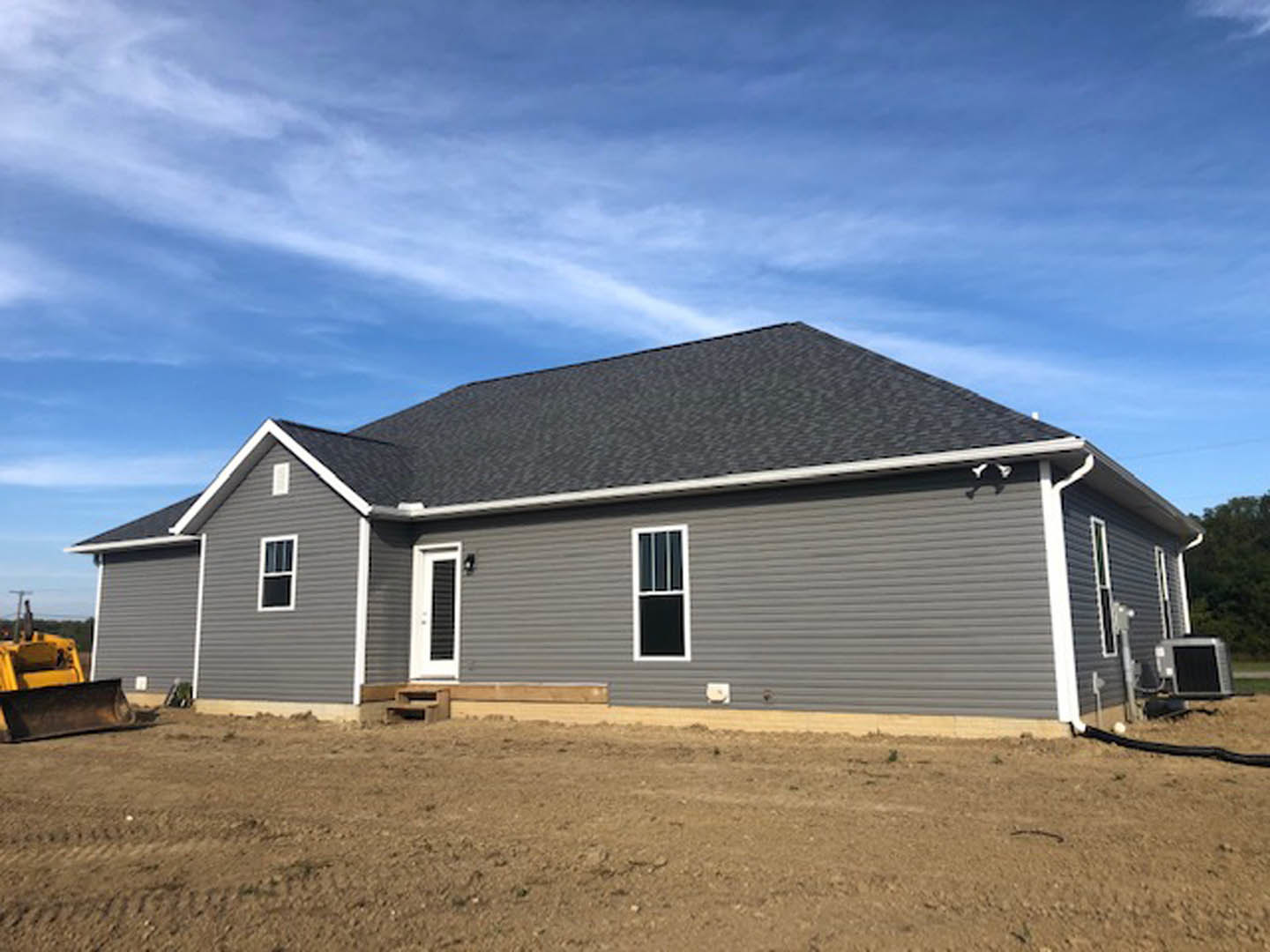 Two-story house under construction with grey roof, white-framed windows, and exposed dirt yard under blue sky