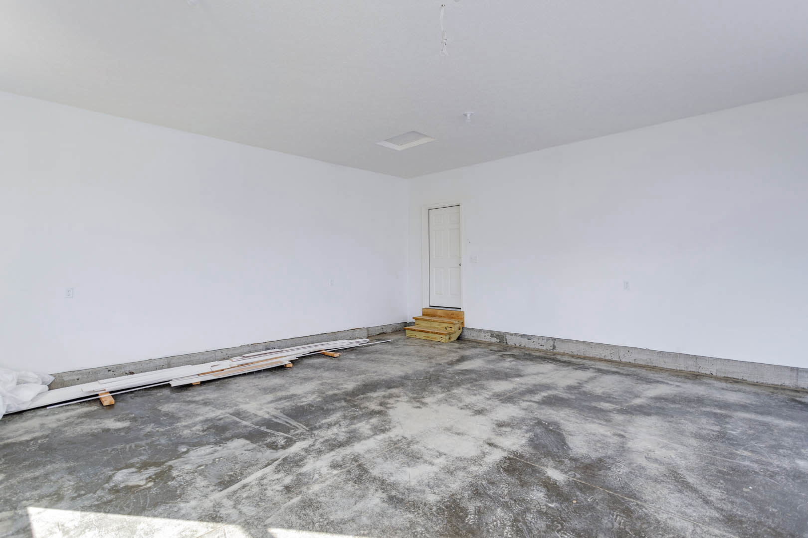White wall with a white door and doorknob, concrete floor with wood planks, wooden steps outside the door, pile of white boards stacked in the room.