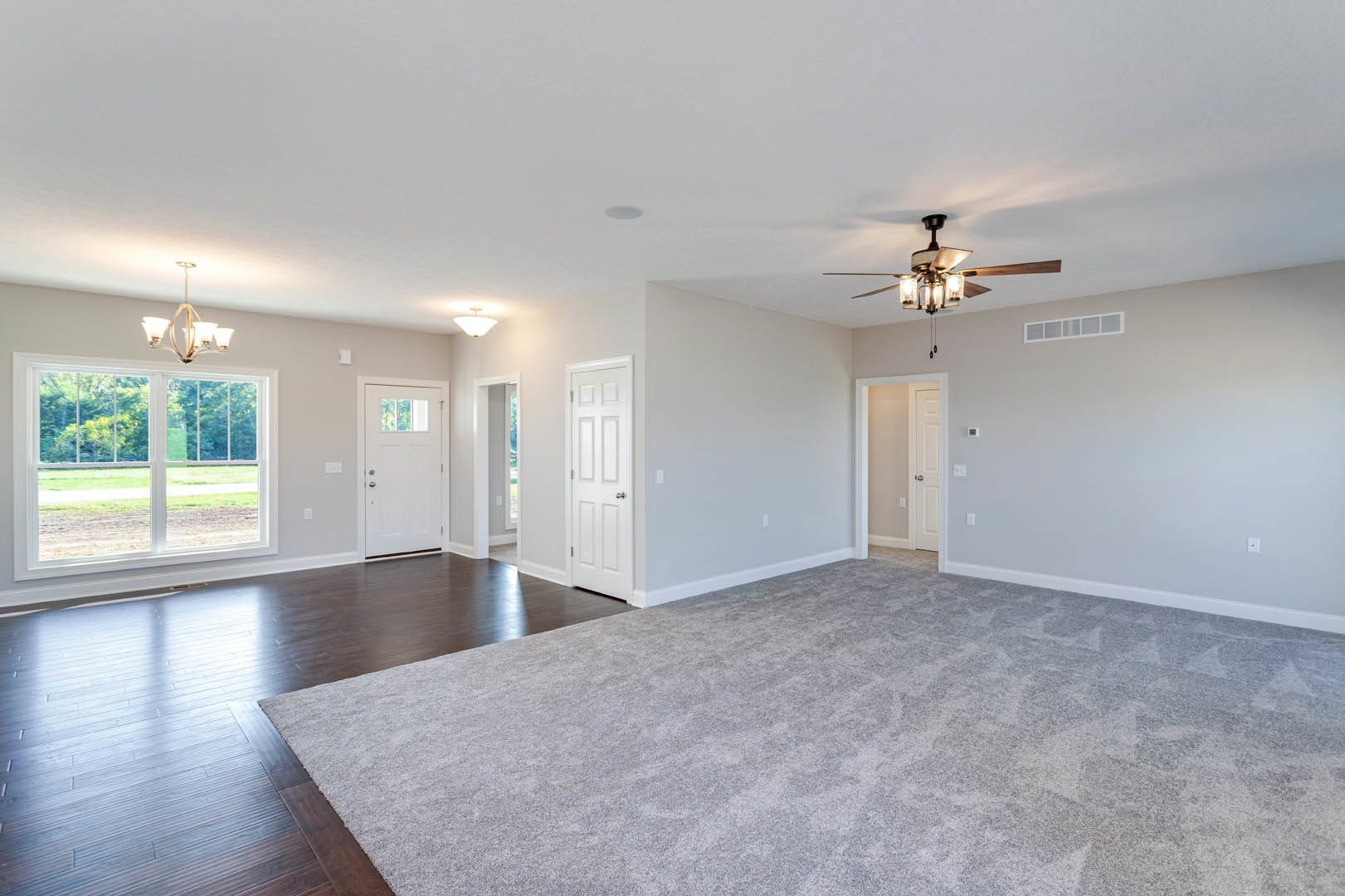 Carpeted room with white walls, ceiling fan, white door with silver handle, window overlooking road and trees, close-up of chandelier