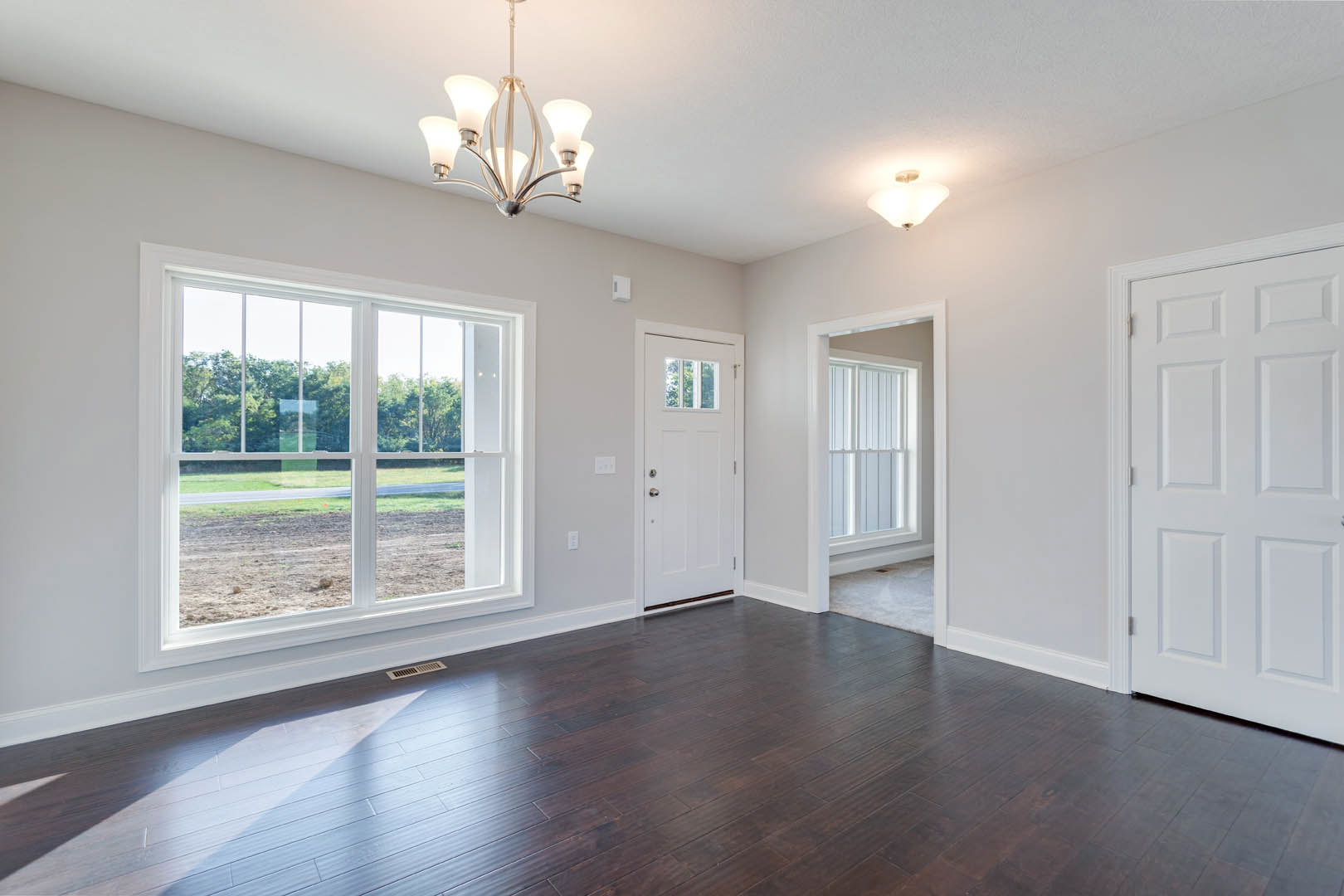 Dark wood flooring with white trim, white door featuring a cross design, large window overlooking a dirt field, five-light ceiling fixture, neutral walls.