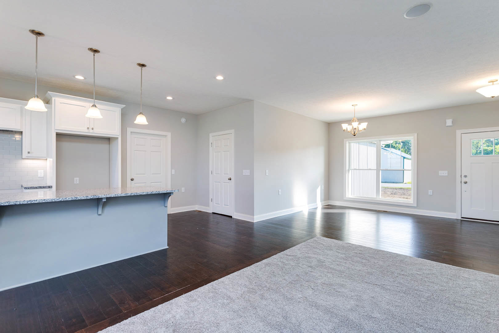Open kitchen with white cabinetry, wood flooring, and a patterned rug; white ceiling with recessed lighting, window with white trim, and adjacent white doors including one with