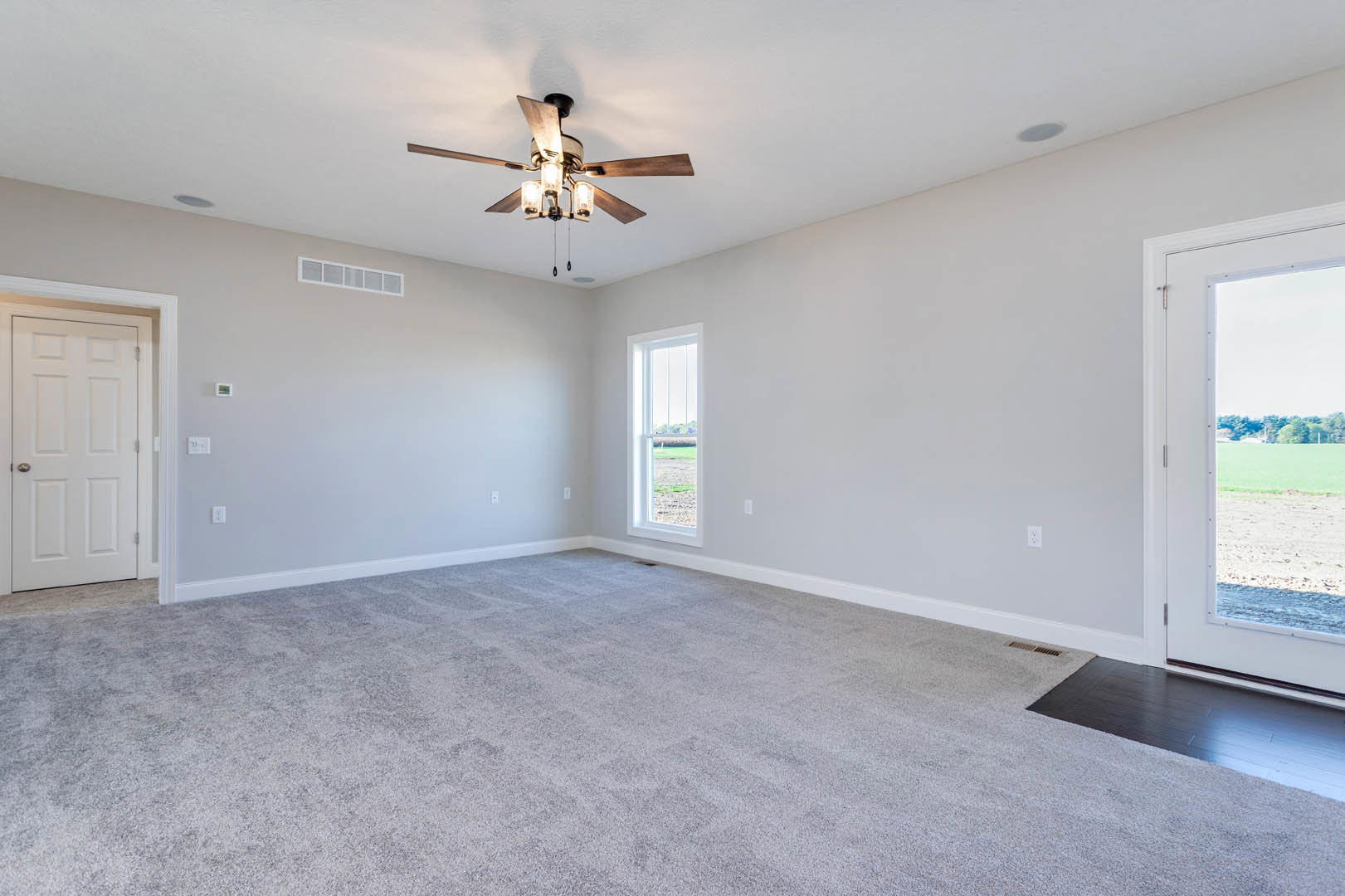 Carpeted bedroom with ceiling fan and light fixture, white door with silver handle, large window overlooking grassy field, neutral walls and trim