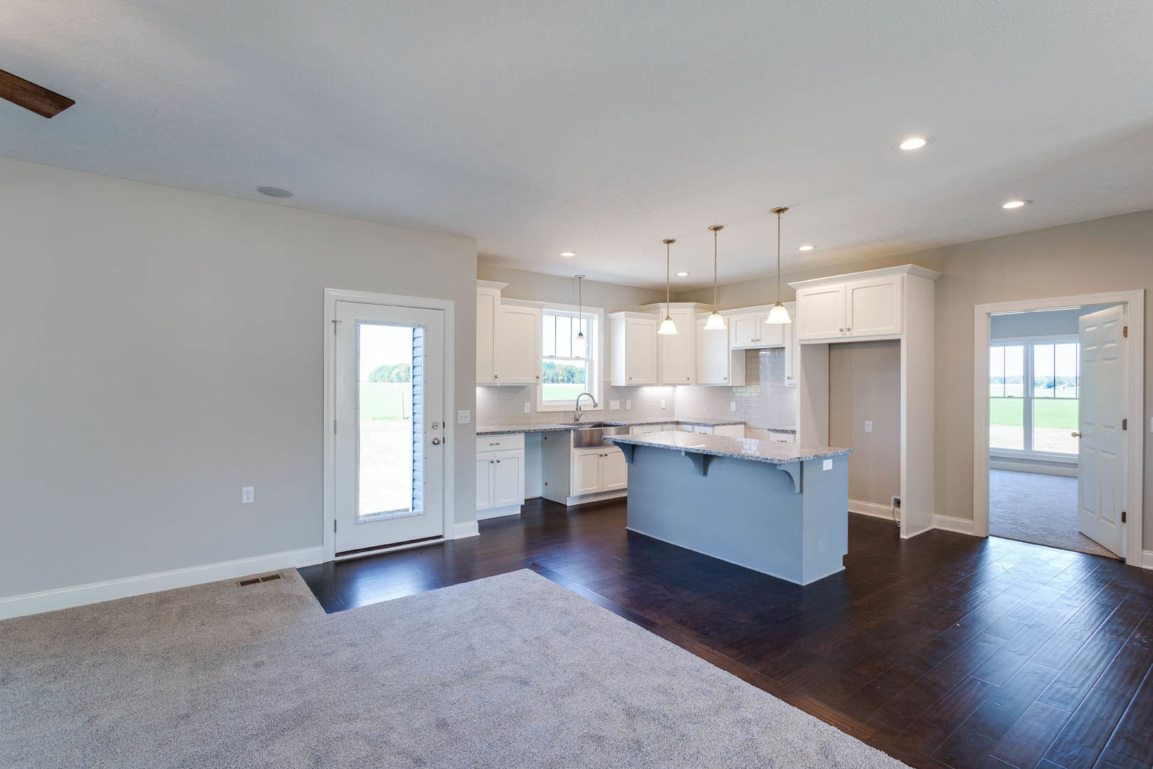 Open concept kitchen and living room featuring hardwood floors, area rug, white cabinetry, countertop with shelf, and two doors—one open to reveal grassy yard and blue sky with