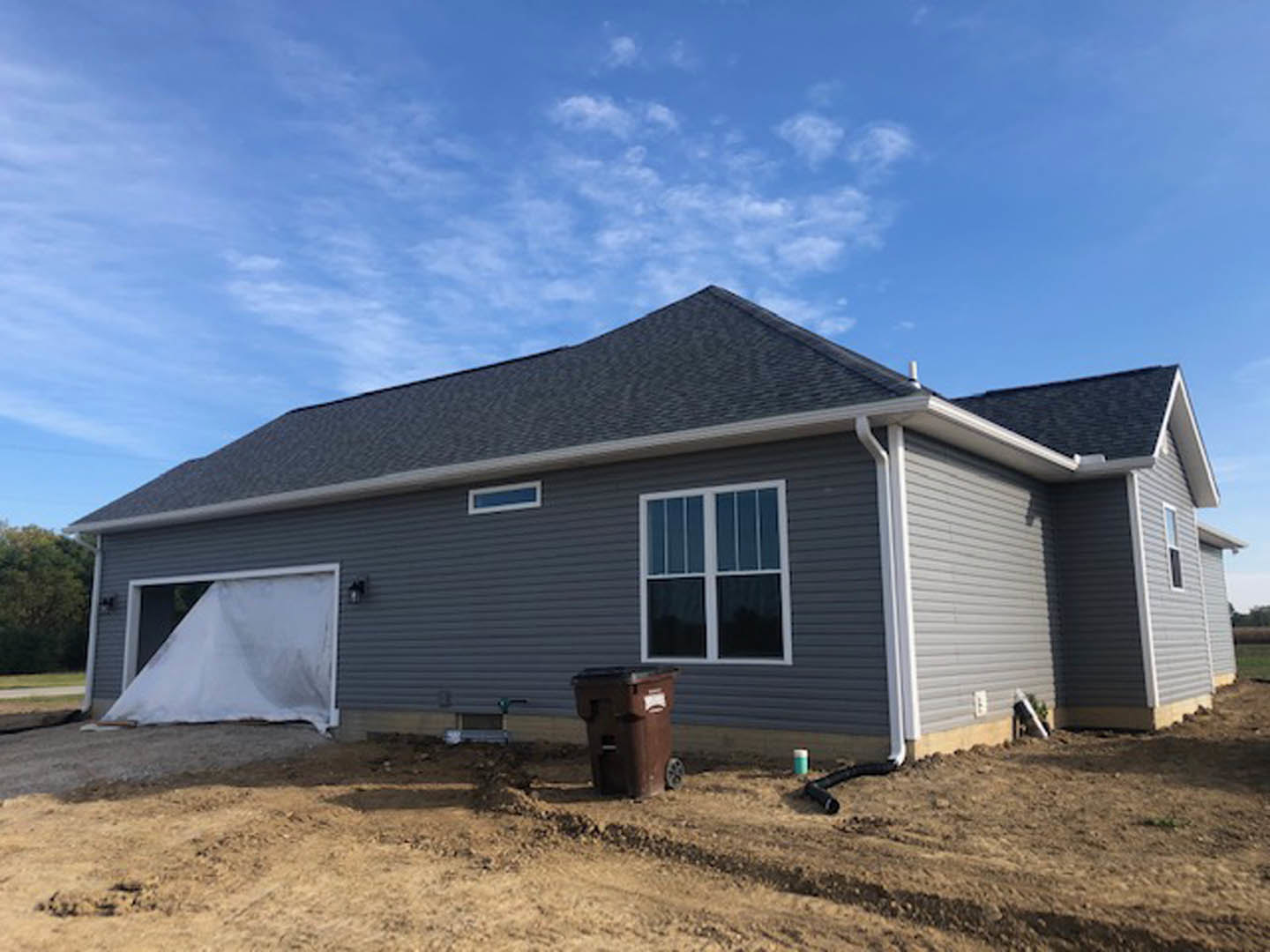 Two-story house under construction with white siding, exposed framing, and a white-framed window; brown wheeled trash can on dirt in front; garage door opening covered with white