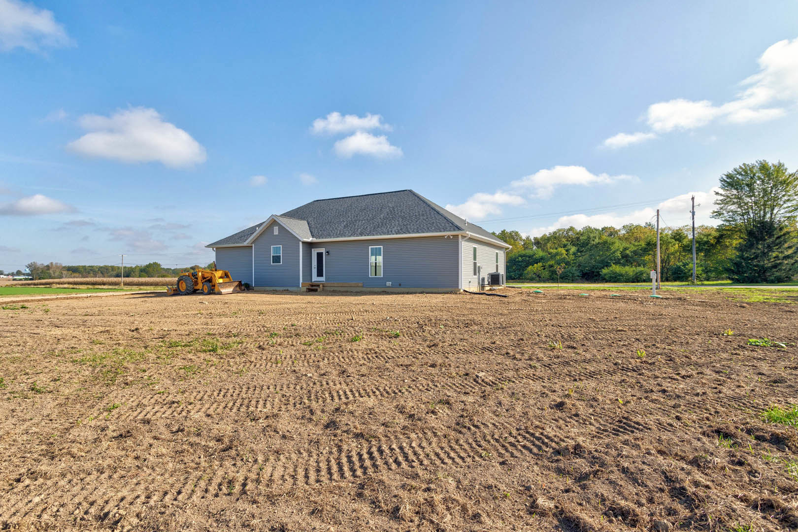 Modern farmhouse with white siding and dark roof sits behind a green tractor parked on bare dirt lot, surrounded by leafy trees under a clear blue sky.
