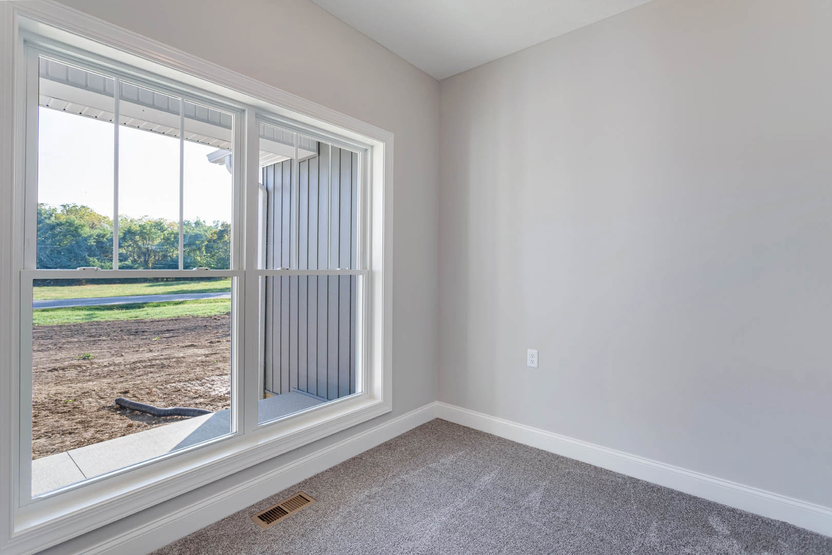 Carpeted room with floor vent, window with blinds overlooking dirt field and tree, neutral walls
