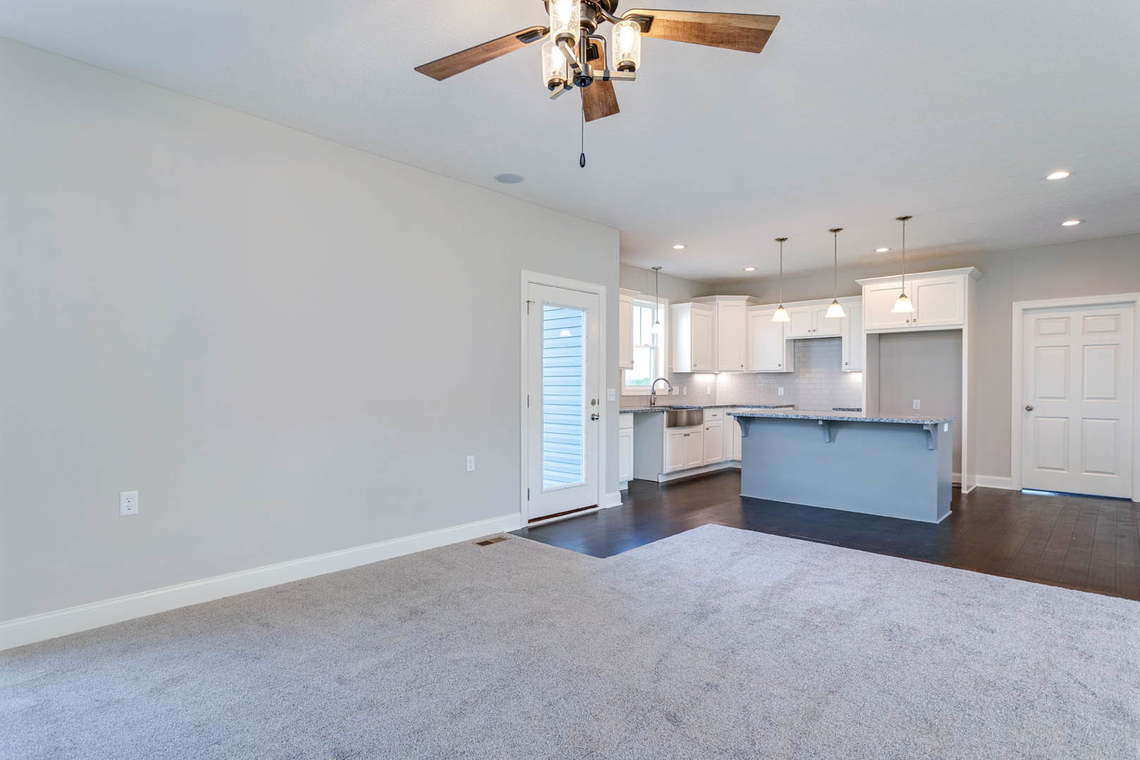 Open concept living room with carpet flooring, adjacent kitchen featuring blue countertops, white cabinetry, and a ceiling fan with lights; white door with window and silver handle