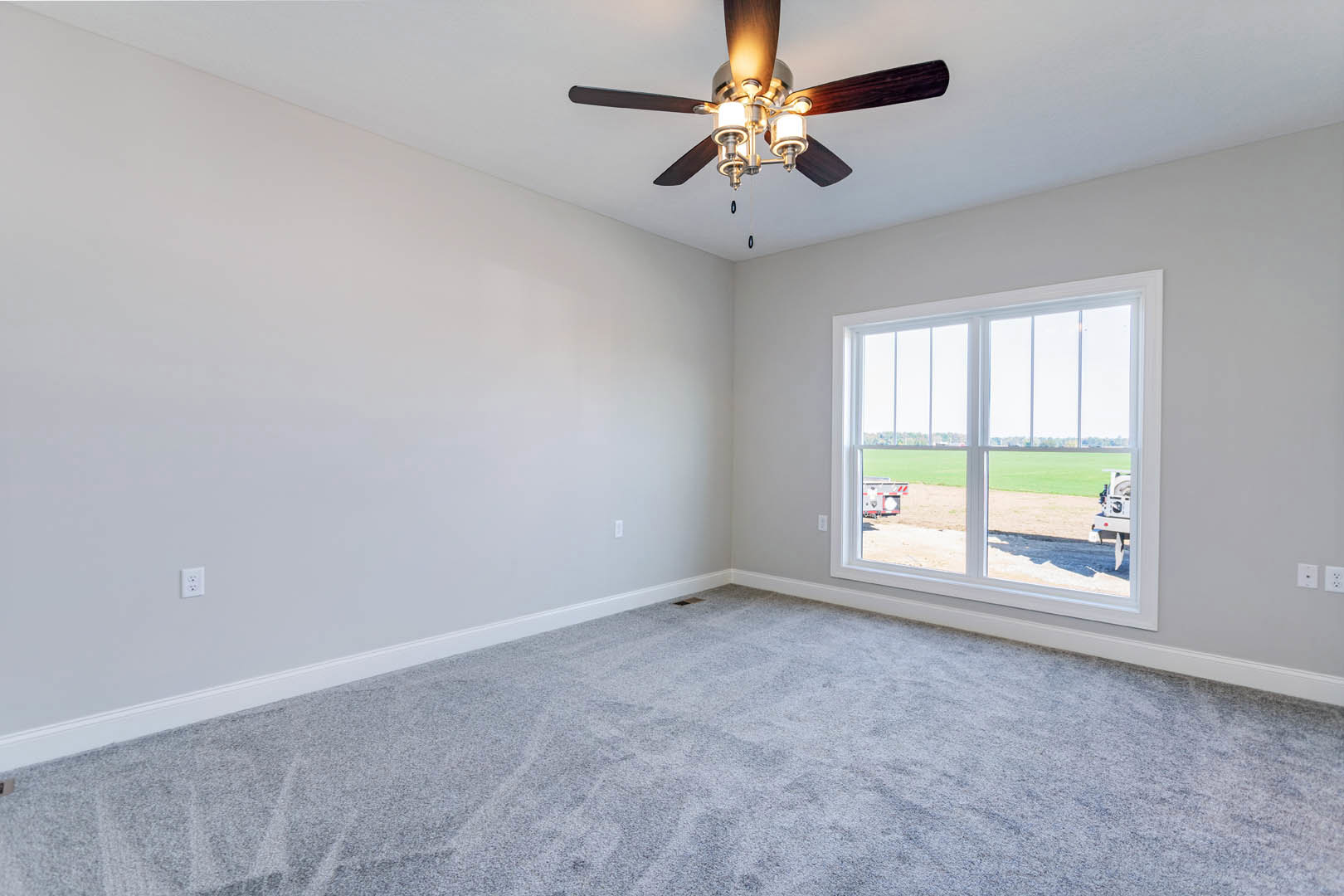 Carpeted room with white walls, ceiling fan with light fixture, large window overlooking grassy field and parked truck, crown molding along ceiling.